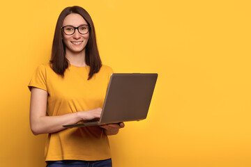 Young cute woman in eyeglasses is smiling happily, holding laptop isolated over yellow background, looking at the camera. Copy space. Promotion concept