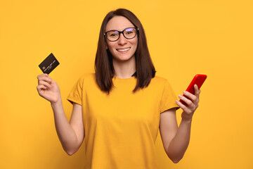Portrait of smiling pretty woman in glasses holding credit card and smartphone isolated over yellow background. Using mobile phone for online banking, shopping and paying bills. Technology concept