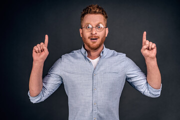 Fototapeta premium Losing words while looking at great business plan. Portrait of amazed and impressed excited redhead man with bristle, gasping, pointing up with raised hands and gazing through glasses at camera