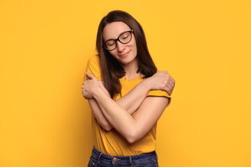 Young beautiful brunette woman in eyeglasses wearing casual yellow t-shirt hugging herself, looks...