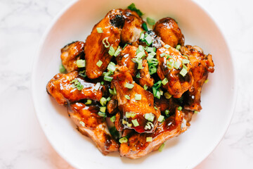 A bowl of sticky and sweet honey and soy sauce glazed chicken wings in a white bowl. They are topped with chopped up scallions. Asian cuisine. 