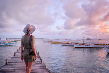 Photography and travel. Young woman in hat holding camera standing on wooden fishing pier with beautiful tropical sea view.