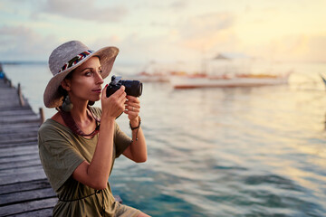 Obraz premium Photography and travel. Young woman in hat holding camera sitting on wooden fishing pier with beautiful tropical sea view.