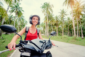 Fototapeta na wymiar Tropical travel and transport. Young beautiful woman in helmet riding scooter on the road with palm trees.
