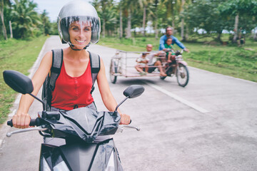 Naklejka premium Tropical travel and transport. Happy young woman in helmet riding scooter on the road with palm trees.