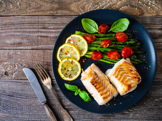 Fish dish - fried cod fillet with asparagus and cherry tomatoes served on black plate on wooden...