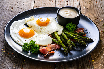 Tasty breakfast. Sunny side up eggs with green asparagus, fried bacon and garlic dip served on black plate on wooden table