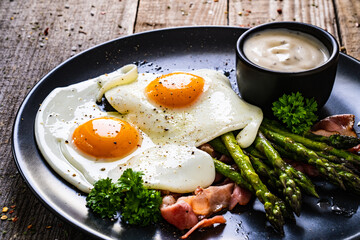 Tasty breakfast. Sunny side up eggs with green asparagus, fried bacon and garlic dip served on black plate on wooden table