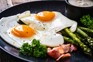 Tasty breakfast. Sunny side up eggs with green asparagus, fried bacon and garlic dip served on black plate on wooden table