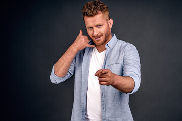 Young attractive guy with red hair in casual shirt, makes phone sign, indicates with index finger...