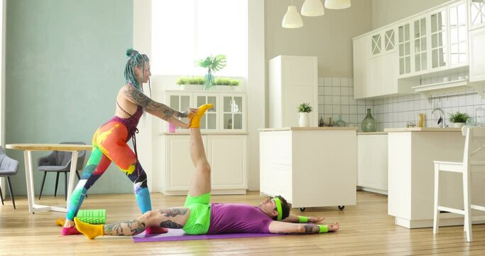 Funny woman with braided hairs stretching man's legs who lying on mat at home. Couple traning together making stretch exercises in kitchen. wife helping her husband. Sport, workout, fitness concept.