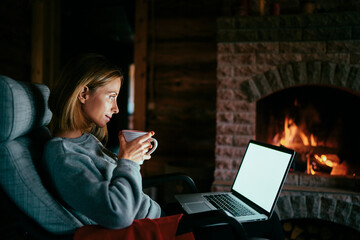 Cozy home. Pretty young woman working on laptop computer near the fireplace. Copy space on the screen.