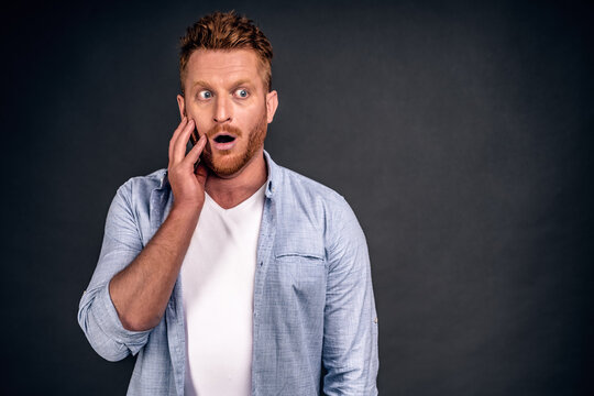 Astounded redhead young handsome male has stubble looks with shocked expression aside, notices something strange on blank grey copy space, area for your advertising content and promotional text.
