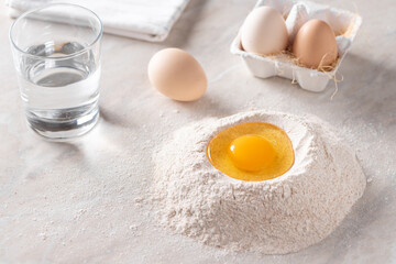 pile of whole grain flour with a raw egg in the middle, a glass of water and eggs on the kitchen table. baking background. place for text. horizontal image. selective focus