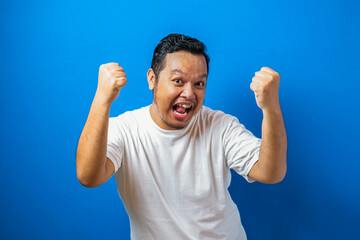Fototapeta premium Portrait of a funny fat Asian man in white t-shirt smiling and dancing happily, joyful expressing celebrating good news victory winning success gesture against blue background