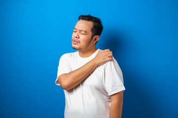 Portrait of good looking young Asian man in white t-shirt having pain on his shoulder, against blue background.