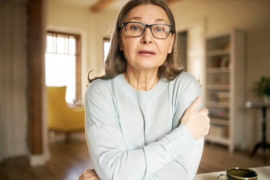 Horizontal shot of gray haired female pensioner in stylish rectangular eyeglasses sitting in living room, looking at camera as if having video conference call. Mature people and age concept