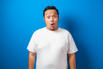 Fat Asian man in white t-shirt shows funny shocked and surprise expression against blue background