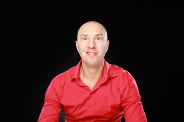 Headshot of bald adult man in red shirt with funny emotions isolated on black background with copy space.