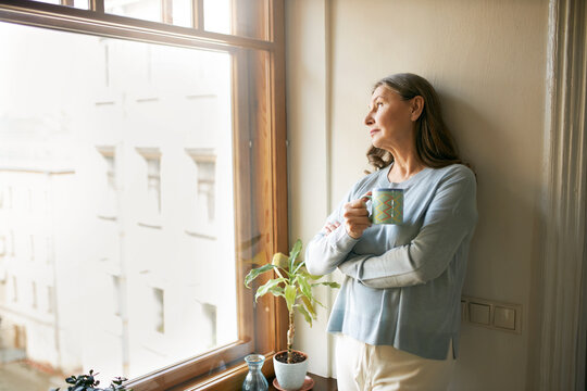 Portrait of thoughtful retired mature woman in casual clothes standing by large window, leaning on white wall, looking outside, drinking tea. People, social distancing and quarantine concept