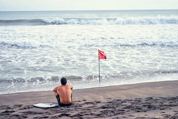 No swimming! Surfer man sitting next to the sea at the beach with red flag.