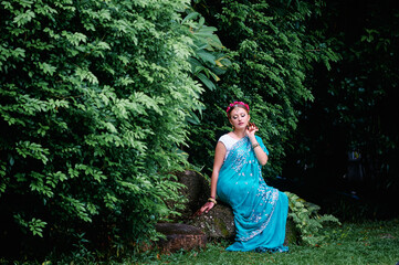 Beautiful young caucasian woman in traditional indian clothing with bridal makeup and jewelry. Bollywood dancer in Sari and henna on hands walking on green garden.