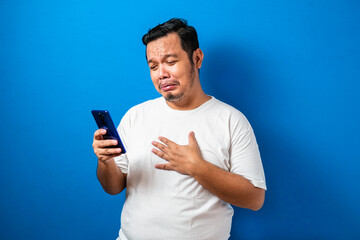 Fat Asian guy wearing a white t-shirt looks sad reading online news from his cellphone. man shows disappointed gesture by wiping tears from his face