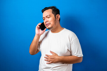 A photo of an asian man in white t-shirt looking sad over the news he received on his smart phone, half upper body studio shot against blue background