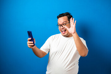 Studio portrait of a young east asian man in white t-shirt making a video call and take selfie, against blue background