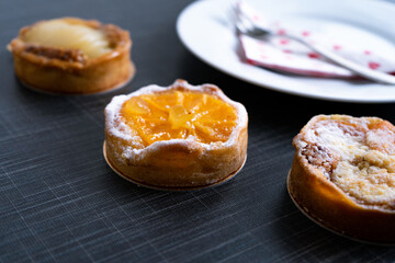 close up of small, french tarts with fruits and powdered sugar placed on dark background with a plate and fork behind