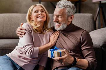 Loving middle aged man making surprise by giving gift in box to his wife celebrating anniversary or birthday at home