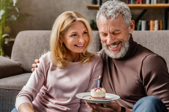 Middle aged couple holding cake celebrating husband's birthday in living room at home
