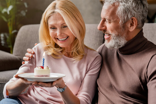 Happy senior couple celebrating anniversary or birthday with cake and candle indoors