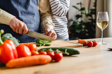 Cropped image of mature wife hugging husband while he cooking vegetable salad at home kitchen