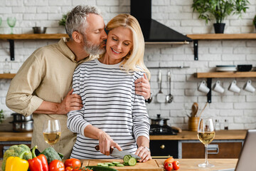 Happy mature couple kissing and hugging in kitchen while cooking