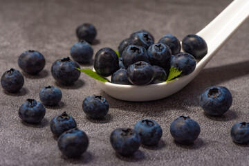 Close-up of a white spoon with juicy fresh blueberries and small green leaves, berries scattered on a dark table.