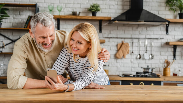 Middle Aged Happy Couple Using Mobile Phone While Standing In Kitchen At Home