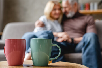 Two coffee cups on table with senior couple relaxing on background on couch