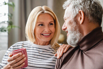 Close up of smiling middle aged woman with her husband relaxing on sofa at home with cup of coffee