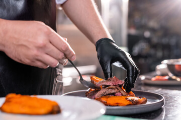 Close-up of unrecognizable chef in glove adding meat steak on plate with vegetables at commercial kitchen