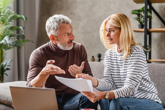 Upset middle aged couple with laptop discussing documents while sitting on couch at home