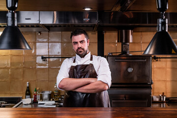 Portrait of serious bearded chef in leather apron standing with crossed arms at restaurant kitchen