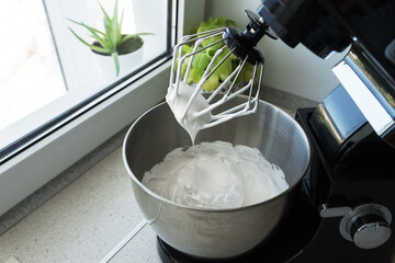 white protein cream on a whisk of a mixer and in a round iron bowl. The process of making sweet protein flakes for dessert. Home cooking concept.