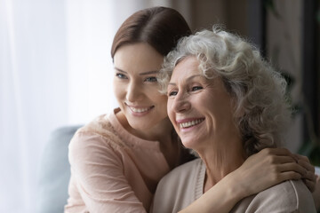 Close up of smiling elderly 60s mother and adult girl child hug look in distance dreaming thinking, happy senior mom and grownup daughter visualize daydream at home together, feel optimistic of future