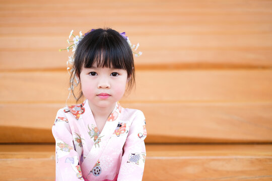 Little asian girl wearing pink traditional japanese dress. Her hair ornaments with flowers. And she sitting on the wooden stairs.
