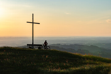 pretty senior woman riding her electric mountain bike in warm dawn sunlight below the summit cross...