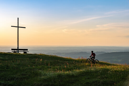 pretty senior woman riding her electric mountain bike in warm dawn sunlight below the summit cross of Salmas hight above Oberstaufen, Allgau Alps, Bavaria Germany 