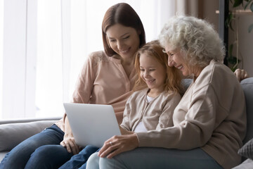 Fototapeta premium Smiling three generations of women relax on sofa in living room watch video movie on laptop, happy little girl with mom and senior grandmother rest on couch at home using modern computer together