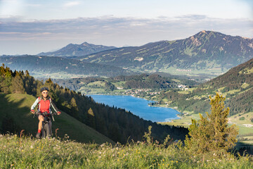 Fototapeta premium pretty senior woman riding her electric mountain bike in warm dawn sunlight heights of Salmas hight above Oberstaufen, with spectacular view on lake Alpsee,Allgau Alps, Bavaria Germany 