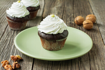 Chocolate cupcake topped with whipped cream, decorated with button-shaped candies. Walnut kernels are lying in front of the plate; two cupcakes and walnut shells are visible in a blurry background.